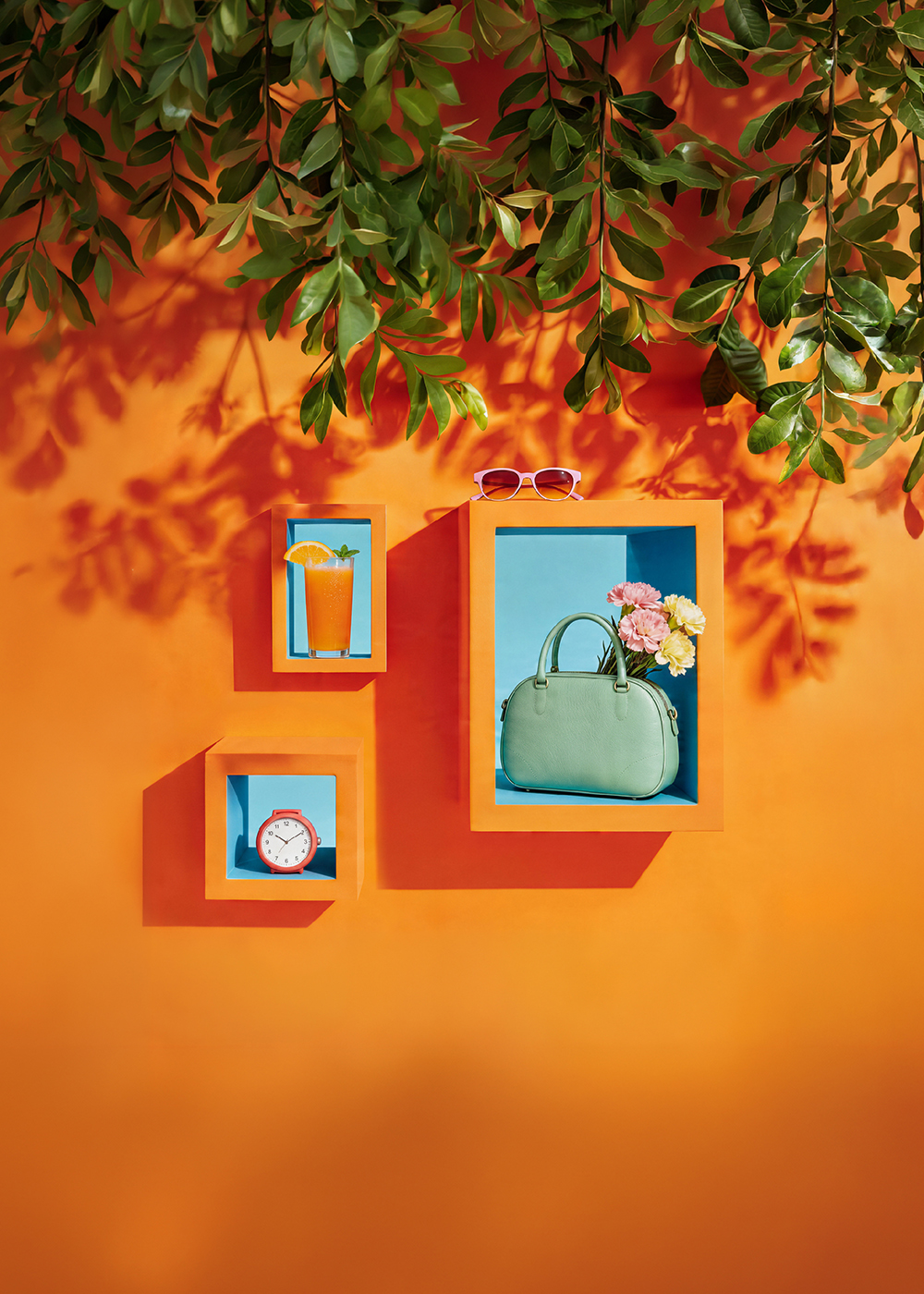 Kolorowa grafika promocyjna: na pomarańczowym tle widoczne są dekoracyjne półki z letnimi elementami – szklanka z napojem i plasterkiem pomarańczy, zielona torebka z kwiatami, zegar oraz okulary przeciwsłoneczne. Grafika nawiązuje do majówki i godzin otwarcia galerii.