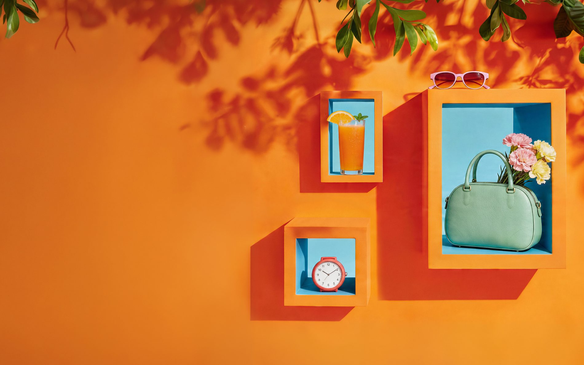 Kolorowa grafika promocyjna: na pomarańczowym tle widoczne są dekoracyjne półki z letnimi elementami – szklanka z napojem i plasterkiem pomarańczy, zielona torebka z kwiatami, zegar oraz okulary przeciwsłoneczne. Grafika nawiązuje do majówki i godzin otwarcia galerii.