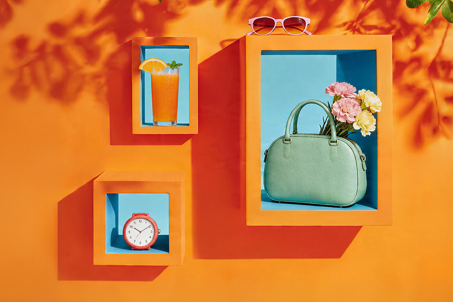 Kolorowa grafika promocyjna: na pomarańczowym tle widoczne są dekoracyjne półki z letnimi elementami – szklanka z napojem i plasterkiem pomarańczy, zielona torebka z kwiatami, zegar oraz okulary przeciwsłoneczne. Grafika nawiązuje do majówki i godzin otwarcia galerii.