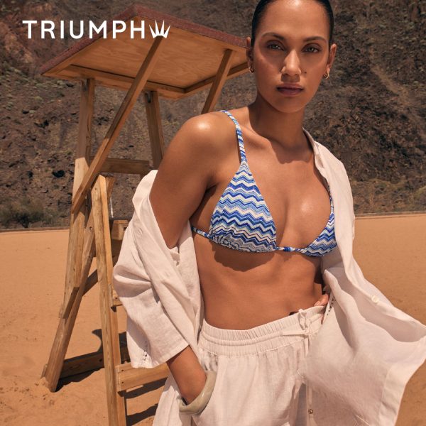 Zdjęcie wizerunkowe marki Triumph prezentujące kolekcję strojów kąpielowych. Modelka pozuje na piaszczystej plaży w dwuczęściowym kostiumie z niebiesko-białym wzorem zygzakowatym, narzuconej białej koszuli oraz białych lnianych spodniach. W tle widoczna drewniana wieża ratownicza oraz górzysty krajobraz. W lewym górnym rogu widnieje białe logo Triumph.
