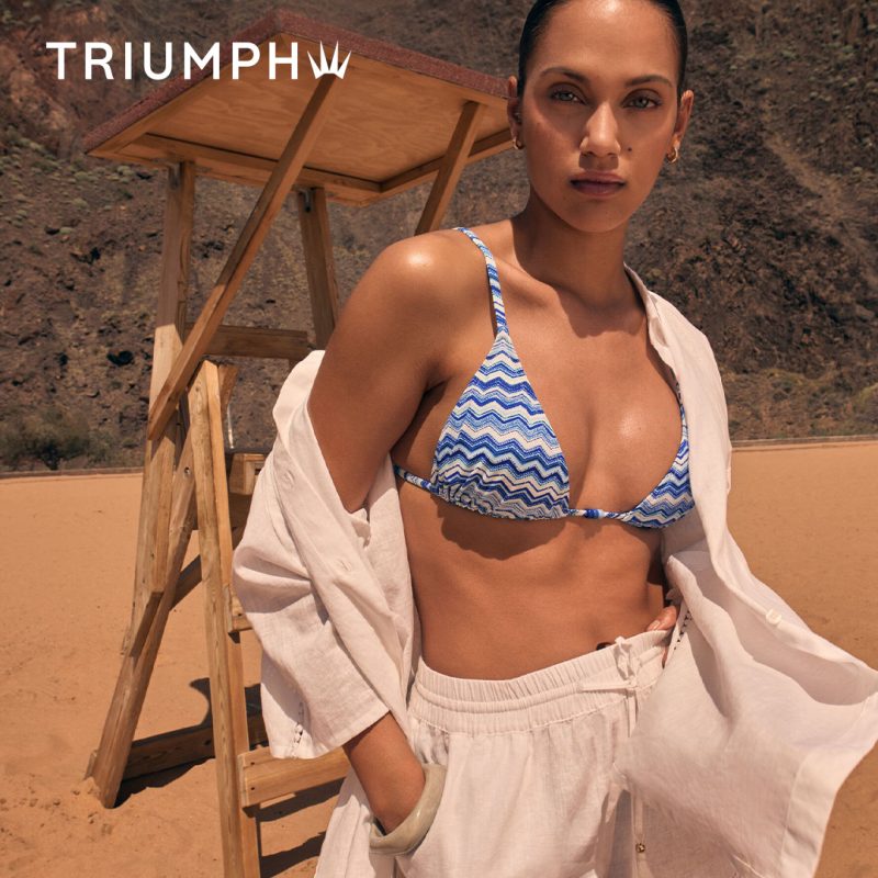Zdjęcie wizerunkowe marki Triumph prezentujące kolekcję strojów kąpielowych. Modelka pozuje na piaszczystej plaży w dwuczęściowym kostiumie z niebiesko-białym wzorem zygzakowatym, narzuconej białej koszuli oraz białych lnianych spodniach. W tle widoczna drewniana wieża ratownicza oraz górzysty krajobraz. W lewym górnym rogu widnieje białe logo Triumph.