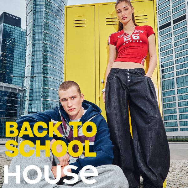 Baner promocyjny House z kampanii Back to School. Para modeli pozuje na tle nowoczesnych wieżowców i żółtej szafki szkolnej. Kobieta stoi ubrana w czerwoną, obcisłą koszulkę z nadrukiem „DIVISION 26” i szerokie, ciemne dżinsy. Mężczyzna siedzi na pierwszym planie, ubrany w granatową bluzę z kapturem z grafiką flagi Union Jack i szare spodnie dresowe. Po lewej stronie duży, żółty napis „BACK TO SCHOOL” i pod nim białe logo „HOUSE”.