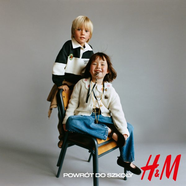Zdjęcie promocyjne marki H&M z kampanii „POWRÓT DO SZKOŁY” na szarym tle. Przedstawia dwoje dzieci: uśmiechniętą dziewczynkę z gwizdkiem w ustach, siedzącą ze skrzyżowanymi nogami na drewnianym krześle, ubraną w jasny kardigan i dżinsy, oraz stojącego za nią chłopca w czarno-białej bluzie polo. W prawym dolnym rogu umieszczono czerwone logo „H&M”, a obok niego biały napis „POWRÓT DO SZKOŁY”.