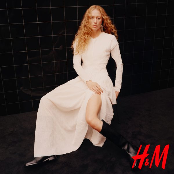 Baner wizerunkowy marki H&M przedstawiający siedzącą na krześle kobietę z długimi, kręconymi rudymi włosami. Modelka ubrana jest w długą, białą sukienkę z rozcięciem na nogę oraz czarne kozaki z wysoką cholewką. Tło tworzy ściana z czarnych, błyszczących kafelków, a w prawym dolnym rogu umieszczono czerwone logo „H&M”.