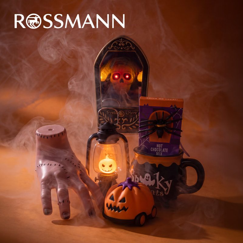 Baner promocyjny Rossmanna w mrocznym, dymnym klimacie Halloween, przedstawiający dekoracje oraz gadżety: sztuczną dłoń ze szwami, świecącą czaskę w gotyckiej ramce, lampion z dynią, kubek z napisem „Spooky”, mieszankę do gorącej czekolady z pająkiem oraz samochodzik w kształcie dyni. W lewym górnym rogu umieszczono białe logo „ROSSMANN”.